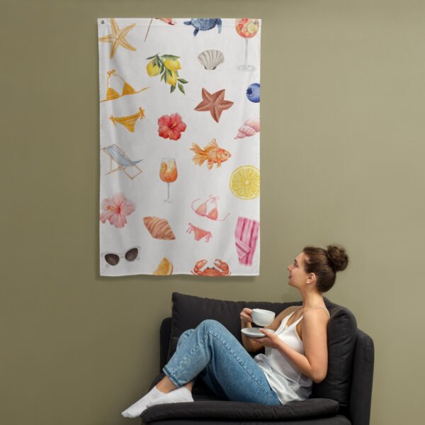 A woman sits on a dark couch holding a cup and saucer, gazing at the Summer vibes tapestry wall hanging on a beige wall. The tapestry displays colorful beach-themed illustrations such as seashells, flowers, sunglasses, and drinks.