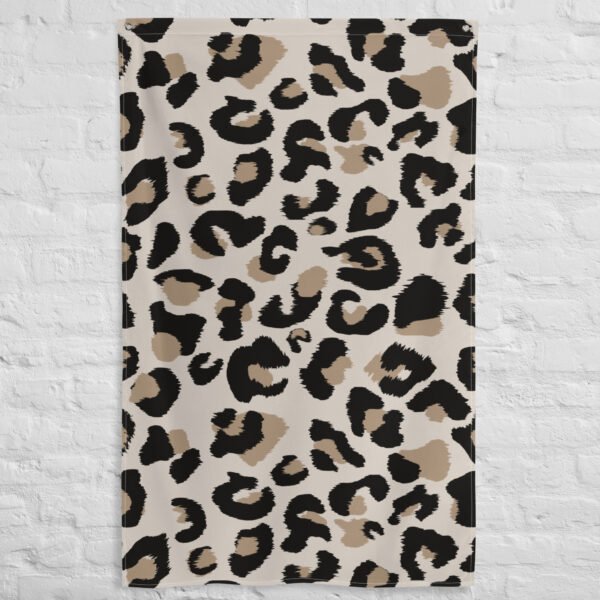 A Leopard print tapestry with a beige background and bold black and brown spots hangs flat against a white textured brick wall.