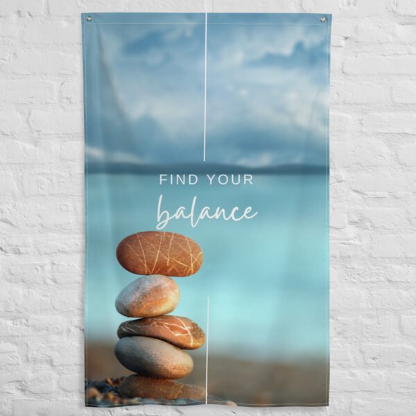 A Find your balance tapestry hangs on a white brick wall, showing stacked smooth stones with a blurred blue sky and water in the background.