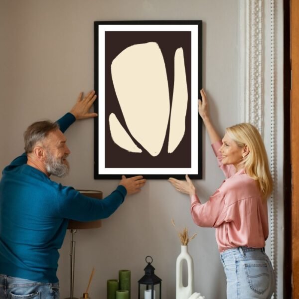 Two individuals hanging a framed abstract artwork featuring a light-colored design on a dark background.