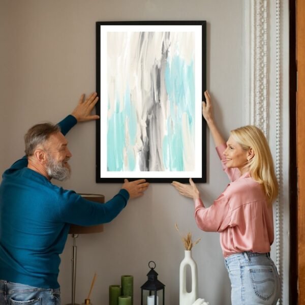 Two individuals placing a framed abstract artwork featuring blue and gray strokes on a wall.