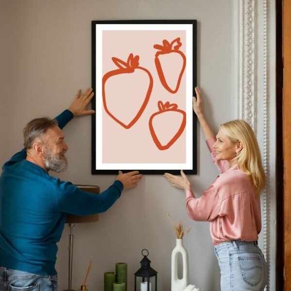 Two individuals hanging a framed artwork featuring three stylized strawberries on a wall.