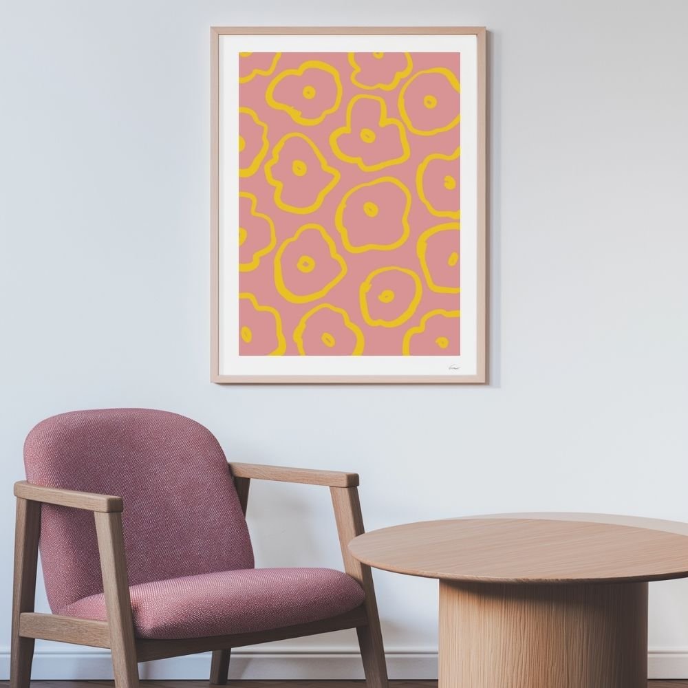 Framed artwork featuring a pink and yellow abstract floral design displayed on a wall next to a chair and table.